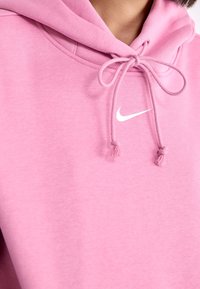 Närbild av en person som bär en rosa huvtröja med en knuten dragsnöre och en liten vit Nike swoosh-logga på bröstet.