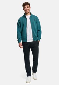 Tyrkis genser med glidelås og høy krage, hvit T-skjorte under, svarte bukser og hvite joggesko, poserer foran en ensfarget hvit bakgrunn.