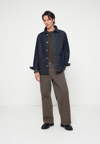 Chaqueta vaquera de color azul oscuro, con botones a presión y dos bolsillos delanteros, combinada con pantalones anchos marrones y zapatos negros.