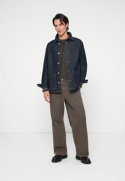 Veste en jean bleu foncé, avec des boutons-pression et deux poches avant, associée à un pantalon large marron et des chaussures noires.