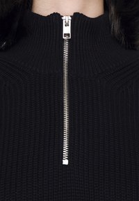 Gros plan d'un pull en maille côtelée noire avec une fermeture éclair en métal argenté partiellement fermée au col montant.