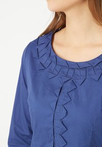 Blusa azul con un diseño texturizado y acentos de tela triangular elevada a lo largo del escote y la tapeta delantera. Acabado de tela suave.