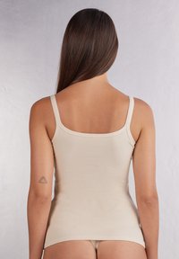 Débardeur beige à fines bretelles, texture lisse, design ajusté et ourlet droit, vu de dos avec une vue partielle du tatouage du modèle.
