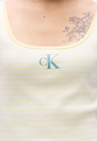 Ριγέ αμάνικη μπλούζα σε λευκό και ανοιχτό κίτρινο, με στρογγυλή λαιμόκοψη και μπλε λογότυπο "CK" στο μπροστινό μέρος, με ραβδωτή υφή.