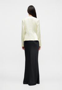 Frau mit langen dunklen Haaren, die steht und einen cremefarbenen Blazer sowie einen langen schwarzen Rock trägt, steht mit dem Rücken zu einem schlichten Hintergrund.