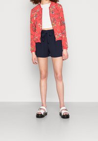 Rød bomberjakke med blomstermønster, hvid crop top, navy shorts med bånd og hvide sandaler med sorte såler. Glatte teksturer og afslappet pasform.