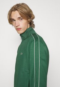 Veste zippée verte avec col monté, rayures blanches sur les côtés et logo brodé. Tissu doux avec une texture lisse. Design ajusté.