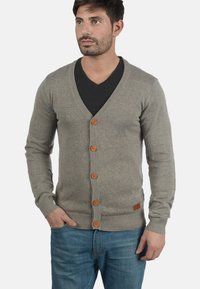 Cardigan grigio a maglia con scollo a V, dotato di bottoni in legno, polsini e orlo a costine. Indossato sopra una maglietta nera e abbinato a jeans blu.