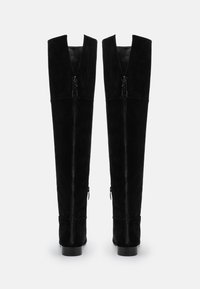 Botas de ante negras por encima de la rodilla con cremalleras verticales en el frente, diseño con cortes en la parte superior y suela plana. Textura suave en toda la superficie.