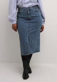 Jupe en denim bleu moyen, avec une fente à l'avant, dotée d'une taille haute et de détails de couture subtils, associée à des bottines noires.