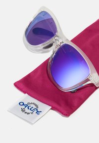 Klare, übergroße Sonnenbrillen mit einem Verlauf in lila Linsen, auf einem magentafarbenen Stoffbeutel platziert, der ein weißes Etikett mit dem Logo "Genuine OAKLEY" trägt.