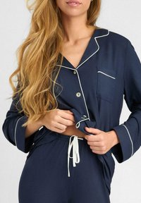 Granatowy komplet piżamowy z miękkiego materiału, z białym wykończeniem, zapinana na guziki bluzka z kieszonką oraz spodnie na sznurku. Luźny krój.