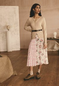 Beige Cardigan über einem floralbedruckten, plissierten Rock in Rosa und Weiß. Braune Wildledersandalen mit Plateau und beigefarbene Socken runden das Outfit ab.