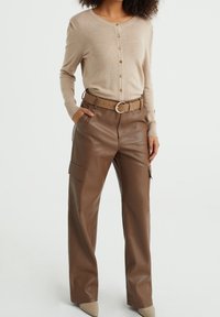 Kvinna klädd i en beige knäppt cardigan instoppad i högmidjade bruna läder cargo-byxor med ett matchande bälte och beige spetsiga skor.