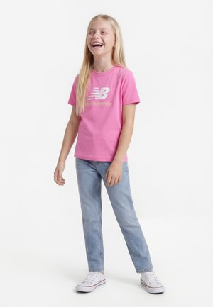 Ragazza giovane in piedi che ride, indossa una maglietta rosa New Balance, jeans azzurri e sneakers bianche su uno sfondo bianco.
