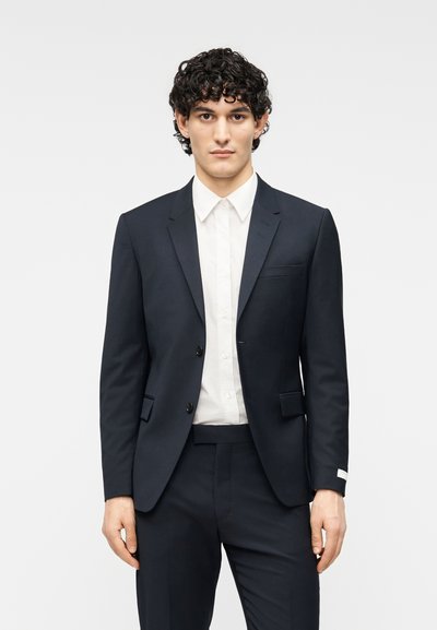 Κοστούμι ναυτικού μπλε με εφαρμοστό σακάκι και ταιριαστό παντελόνι. Διαθέτει γιακά με σχισμή, δύο κουμπιά και τσέπη στο στήθος· συνδυάζεται με λευκό πουκάμισο.