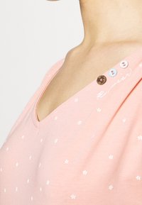 T-shirt in cotone rosa con scollo a V, stampa floreale bianca e bottoni decorativi blu, rosa e marrone sulla spalla.