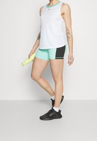Canottiera sportiva bianca con dettagli turchesi, abbinata a pantaloni corti color acquamarina e neri. Sneakers nere e una bottiglia d'acqua gialla completano l'outfit.