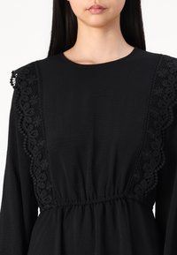 Black dress featuring a textured fabric, round neckline, and long sleeves. Lace detailing along the shoulders and an elastic waistband.