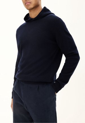 Homme portant un pull à capuche bleu marine et un pantalon bleu foncé assorti, une main dans la poche, sur un fond uni.