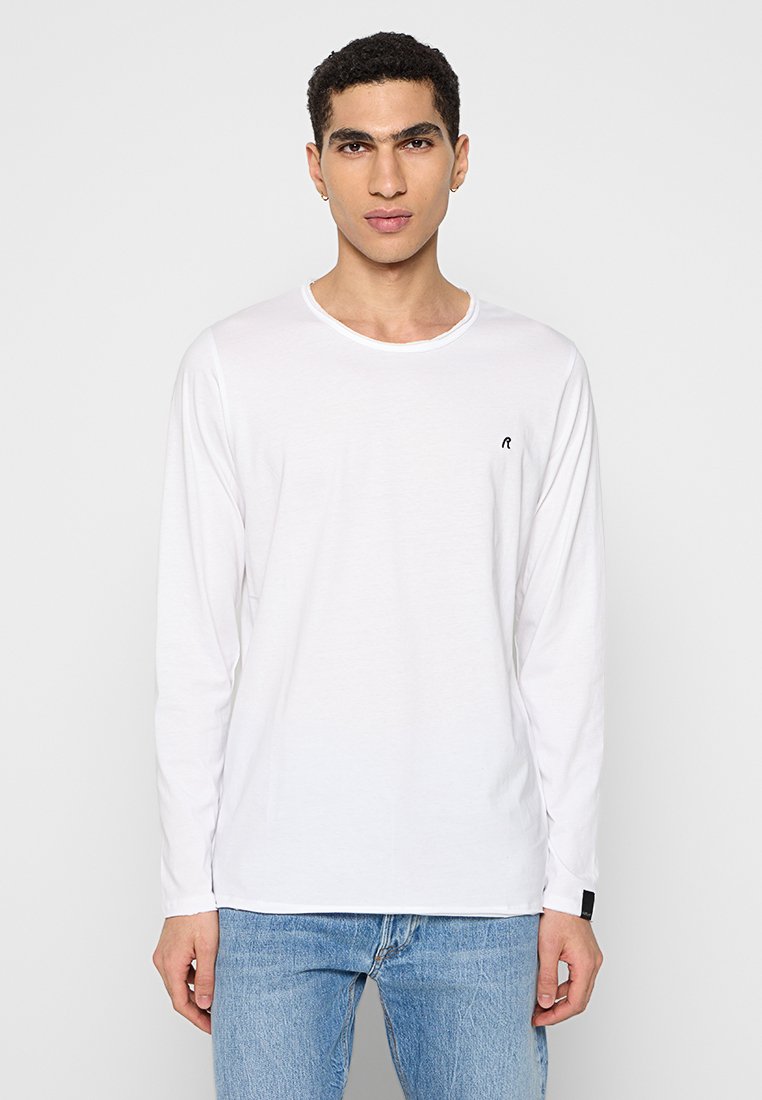 Camiseta de manga larga blanca hecha de algodón, con un cuello redondo y un pequeño logo negro en el lado izquierdo del pecho. Combinada con unos vaqueros azules desgastados.