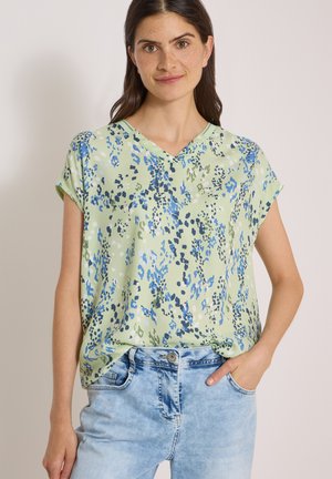 Femme aux longs cheveux foncés portant un haut vert à manches courtes avec un motif abstrait bleu et un jean en denim bleu clair, debout avec une main dans la poche.