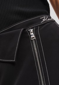 Zwarte rok met een zijrits met zilveren hardware en witte stiksels. De stof heeft een gladde textuur met een gestructureerd ontwerp.