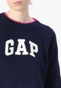 Pulover bleu marin cu guler rotund și finisaj roz. Partea din față afișează litere mari brodate de culoare albă care spun "GAP". Textură moale.