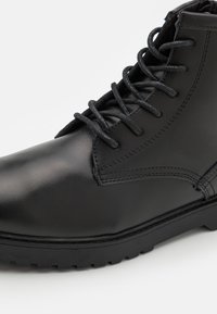 Černé kožené kotníkové boty s hladkým povrchem, kulatou špičkou a černými tkaničkami. Obsahují texturovanou černou gumovou podrážku s vzorem. 