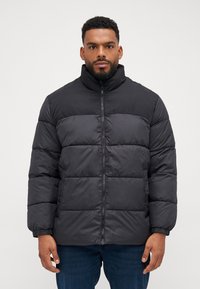 Sort puffet jakke med høj krave, med lynlås lukning, quiltede paneler og elastiske manchetter. Båret over blå jeans.