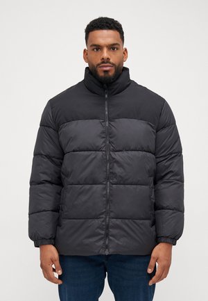 Sort puffet jakke med høj krave, med lynlås lukning, quiltede paneler og elastiske manchetter. Båret over blå jeans.