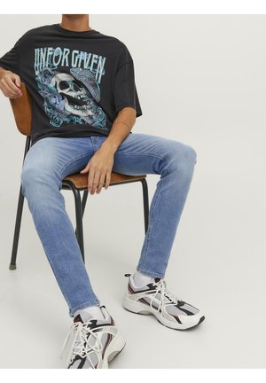 Person sidder på en træstol iført en sort t-shirt med grafik, lyseblå jeans, hvide og sorte sneakers og hvide sokker mod en ensfarvet baggrund.