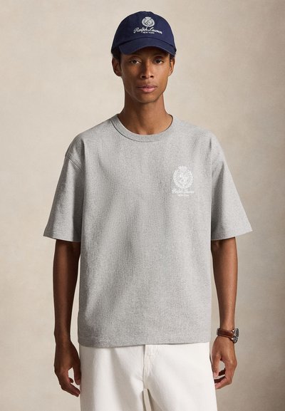 Άνδρας που φορά γκρι μπλουζάκι Ralph Lauren και μπλε σκουφάκι, με λευκό παντελόνι και ρολόι χειρός, στέκεται μπροστά σε μονόχρωμο φόντο.