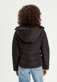 Doudoune noire avec capuche, comportant des panneaux matelassés horizontaux, une fermeture éclair et une coupe décontractée. Portée sur un jean bleu clair.
