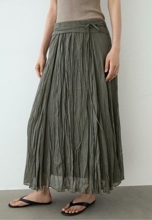 Femme portant une jupe longue olive froissée avec une ceinture nouée, un haut beige sans manches et des tongs noires, debout sur un sol gris.