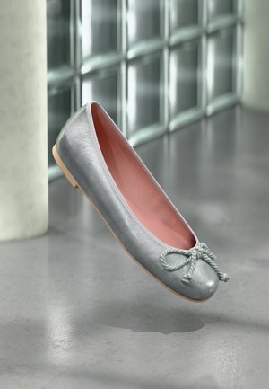 Zilveren ballerina met roze binnenkant en geknoopte strik, zwevend boven een grijs gepolijste vloer nabij een glazen blokmuur.