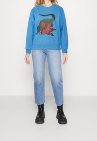 Blå sweatshirt med en grön dinosaurie och grafisk röd blomma, ribbad mudd och nederkant. Matchas med ljusblå raka jeans och svarta stövlar.
