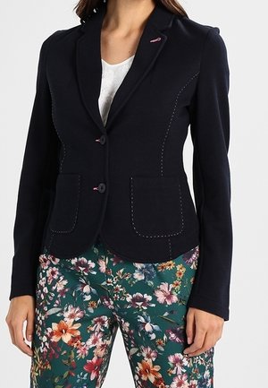 Mujer con una blazer negra ajustada con detalles de costuras rosas sobre una camiseta blanca, combinada con pantalones verdes de estampado floral.