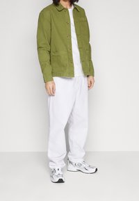 Grön bomullsjacka med krage, framknäppning och sidfickor, i kombination med en vit t-shirt och avslappnade vita byxor. Vita sneakers.