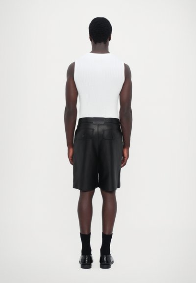 Un débardeur blanc côtelé et un short en cuir noir, avec deux poches arrière, complété par des chaussures et des chaussettes noires. Le modèle est vu de dos.