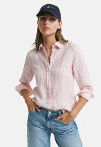 Camicia a righe rosa chiaro e bianca con fit rilassato e maniche arrotolate, abbinata a jeans in denim blu. Cappello nero con logo dell'ancora.