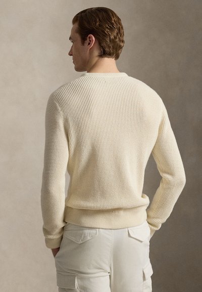 Πλεκτό κρεμ πουλόβερ με υφή, λαιμόκοψη και μανσέτες με ριμπ ύφανση, συνδυασμένο με ανοιχτό μπεζ παντελόνι με πίσω τσέπες.