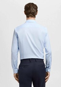 Svetlo modra srajca z dolgimi rokavi in klasičnim ovratnikom, z rahlo teksturo. Oblečena s temnimi hlačami, pogled od zadaj.