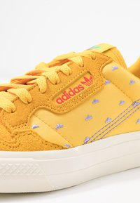 Gula sneakers i mocka och textil, med ett strukturerat mönster, snörningsdesign och en röd adidas-logotyp på sidan.