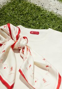 Kremowa bawełniana koszulka z szóstokątną czerwoną metką. Na koszulce leży biała jedwabna chusta z czerwonymi akcentami i wzorami. Tło stanowi zielona trawa.