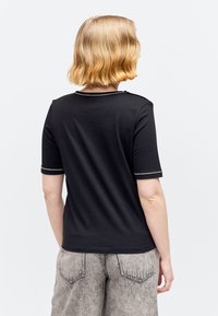 Personne aux cheveux blonds courts portant un t-shirt noir à manches courtes avec des bordures blanches et un jean gris clair délavé à l'acide, vu de dos.