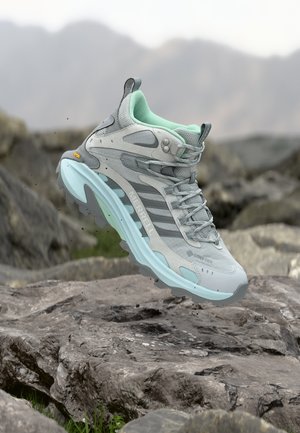 Szürke és menta színű Merrell túracipő Vibram talppal, sziklás terep fölött lebegve, elmosódott hegyekkel a háttérben.