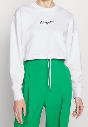 Osoba ubrana w białą krótką bluzę z logo "Hugo" oraz jasne zielone, plisowane spodnie z wysokim stanem, ręce swobodnie opuszczone wzdłuż ciała.