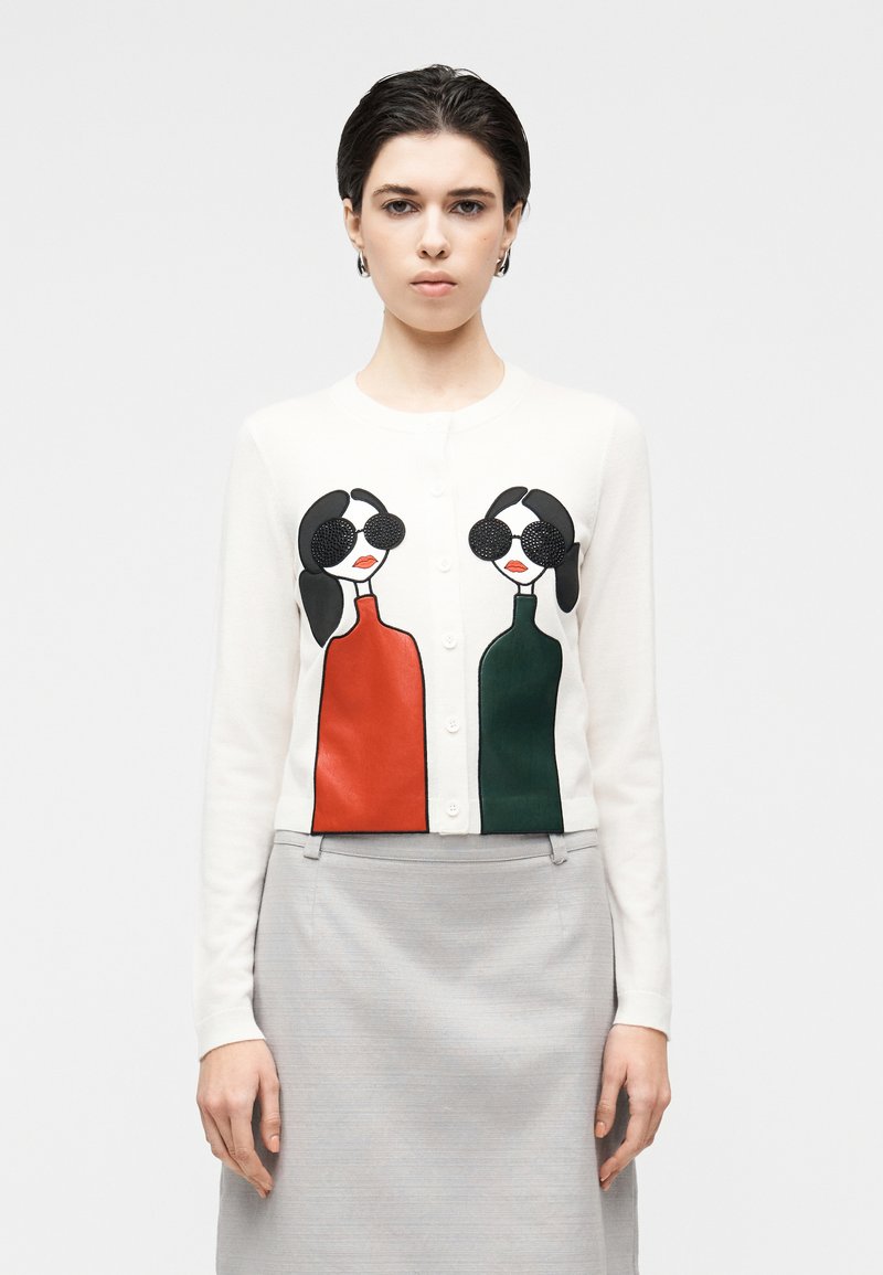 Bela jopica na gumbe, z dvema ilustriranima figurama v rdeči in zeleni majici. Gladek tekstura z vezenimi poudarki.