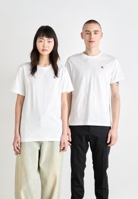 White cotton t-shirts with short sleeves, featuring a small black logo on the left chest; paired with light green and black pants.